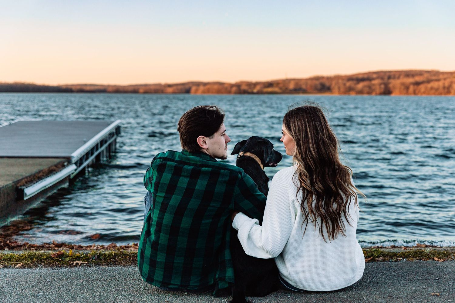 Two people sit together with their dog on a dock overlooking a serene lake during sunset.