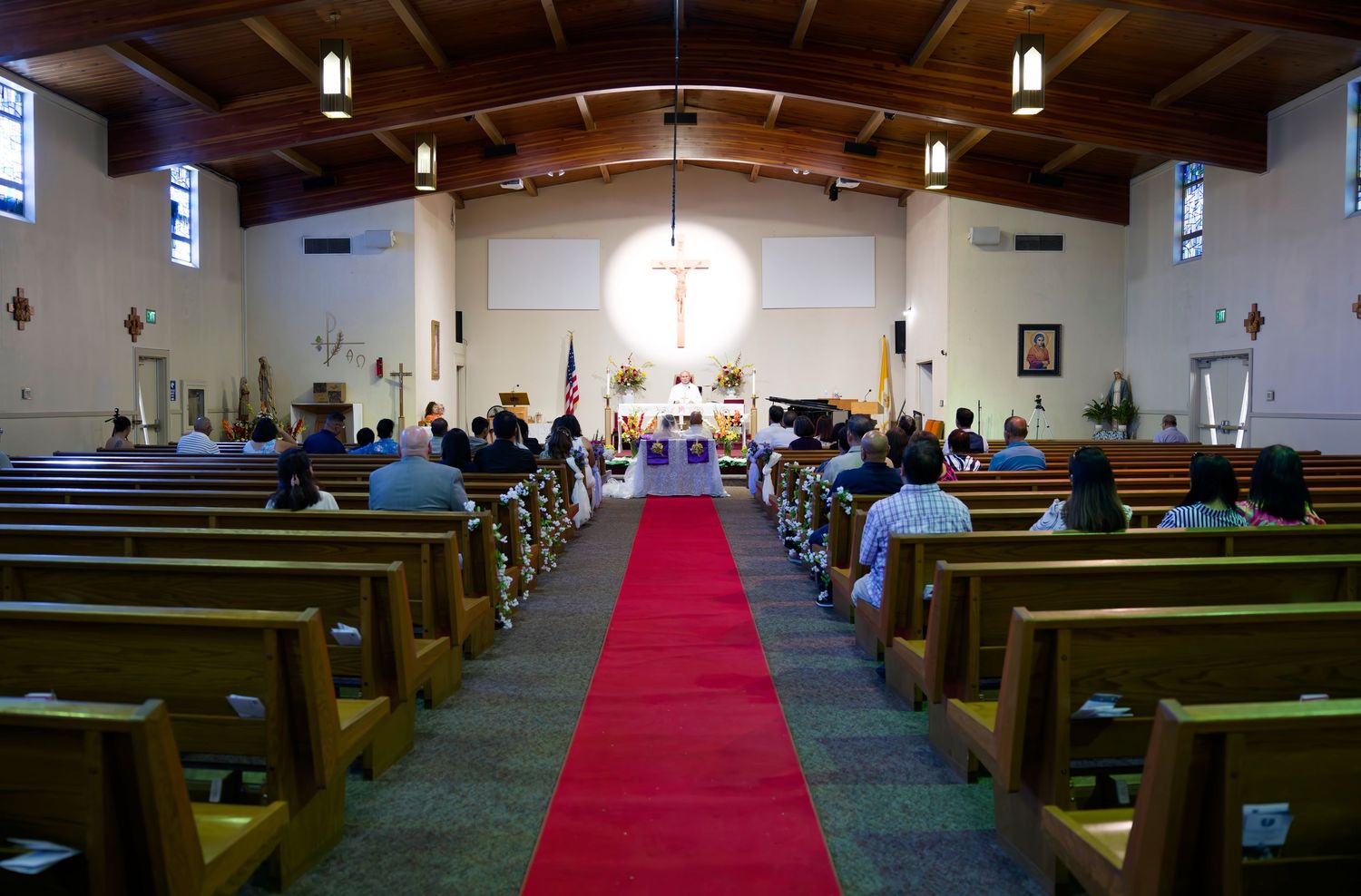 Church interior with wooden pews, red carpet aisle, and illuminated cross at altar during a wedding ceremony.