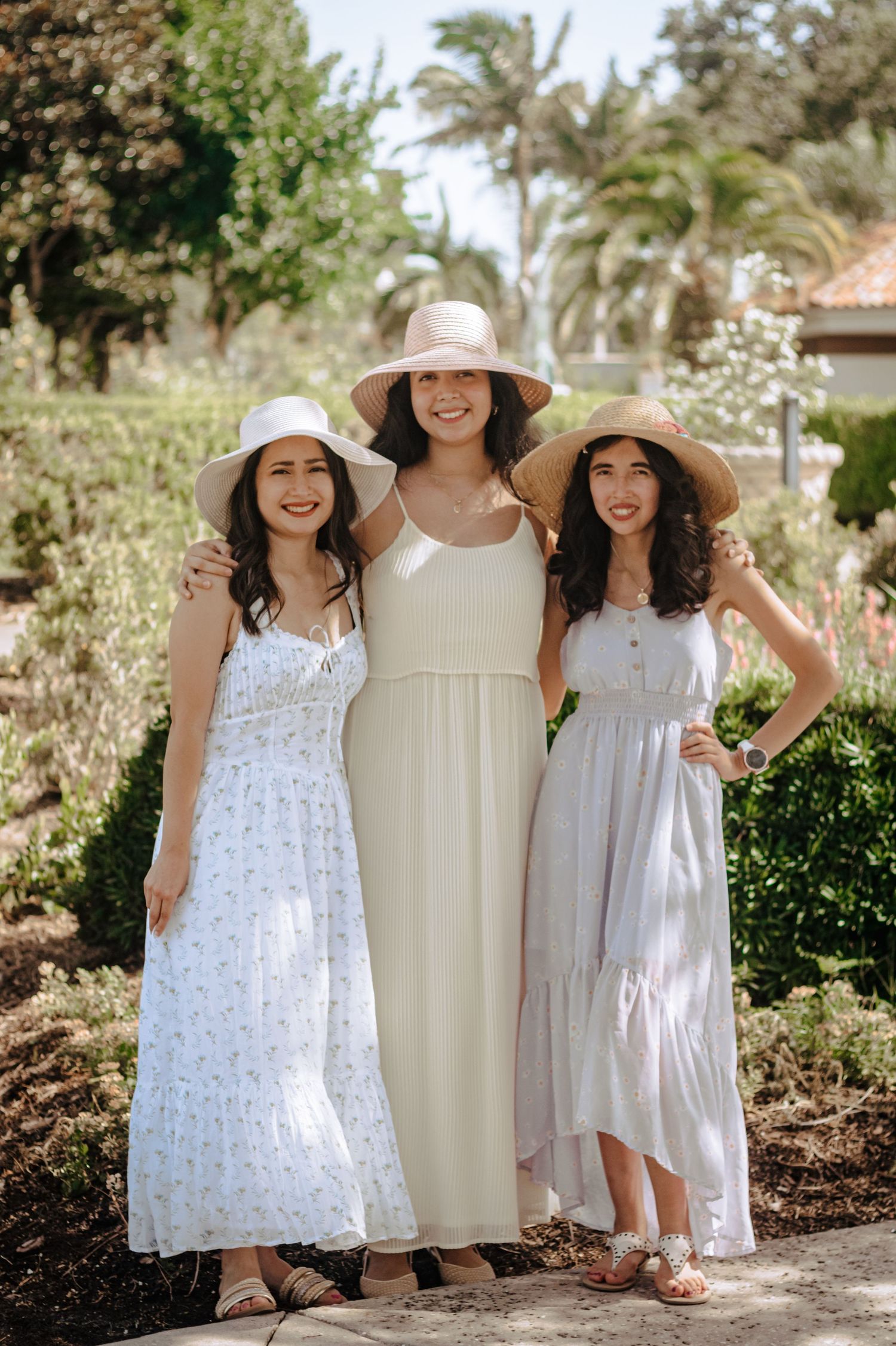 Three smiling women in white summer dresses and straw hats posing together in a sunny garden.