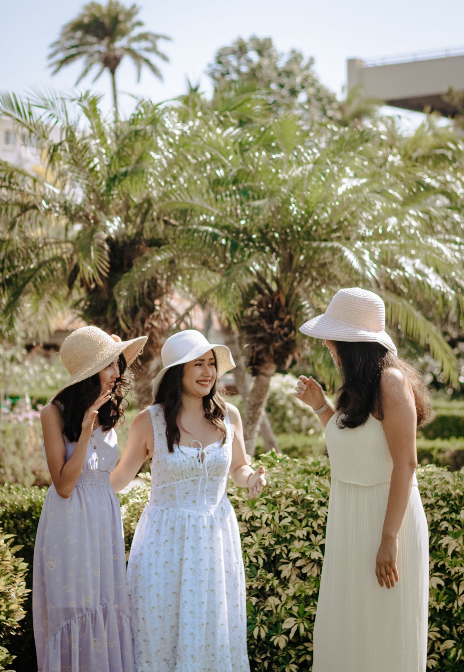 Three young women in white summer dresses and sun hats chatting in a sunny tropical garden.