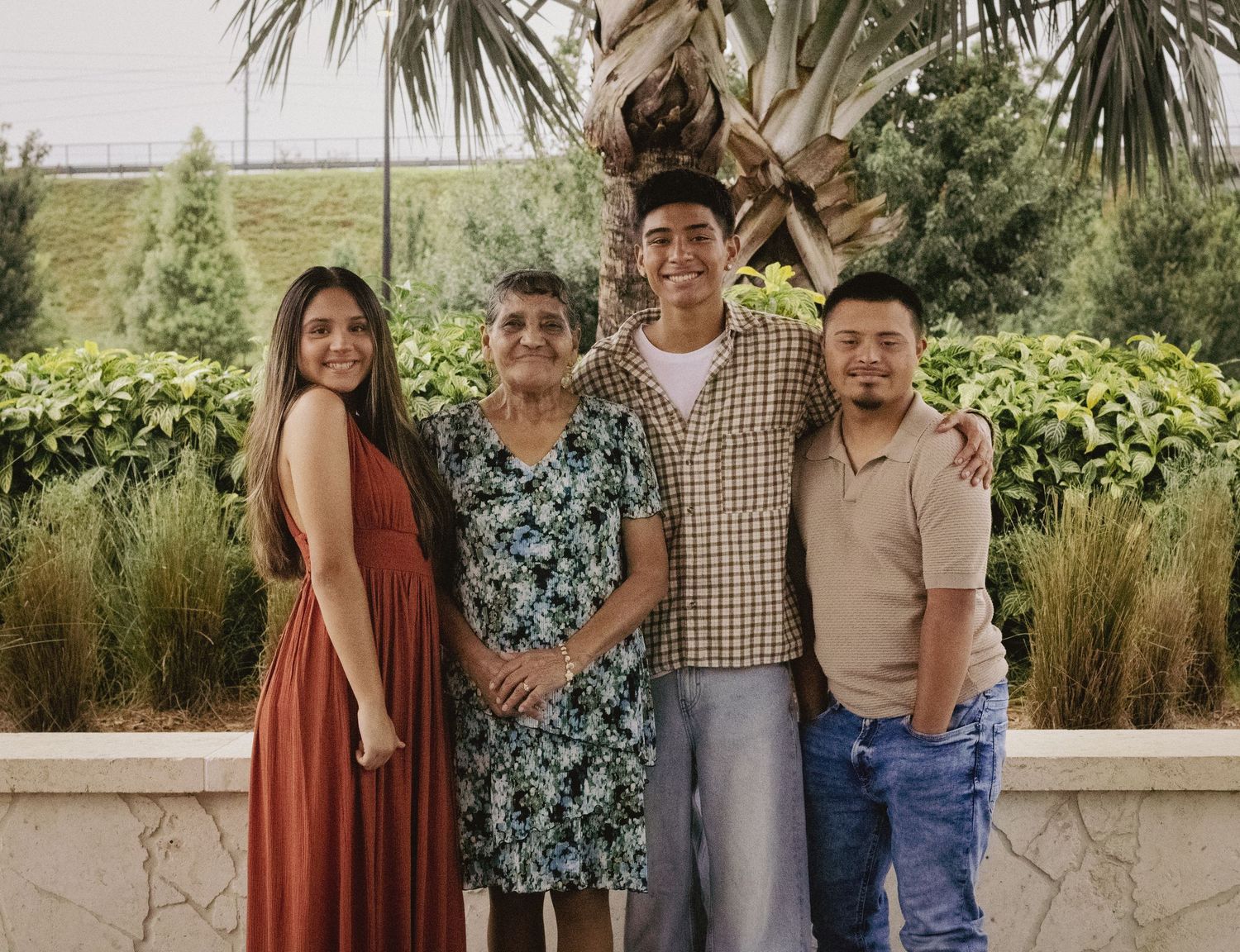 Family of four posing together outdoors near tropical plants, featuring an elderly woman and three younger adults.