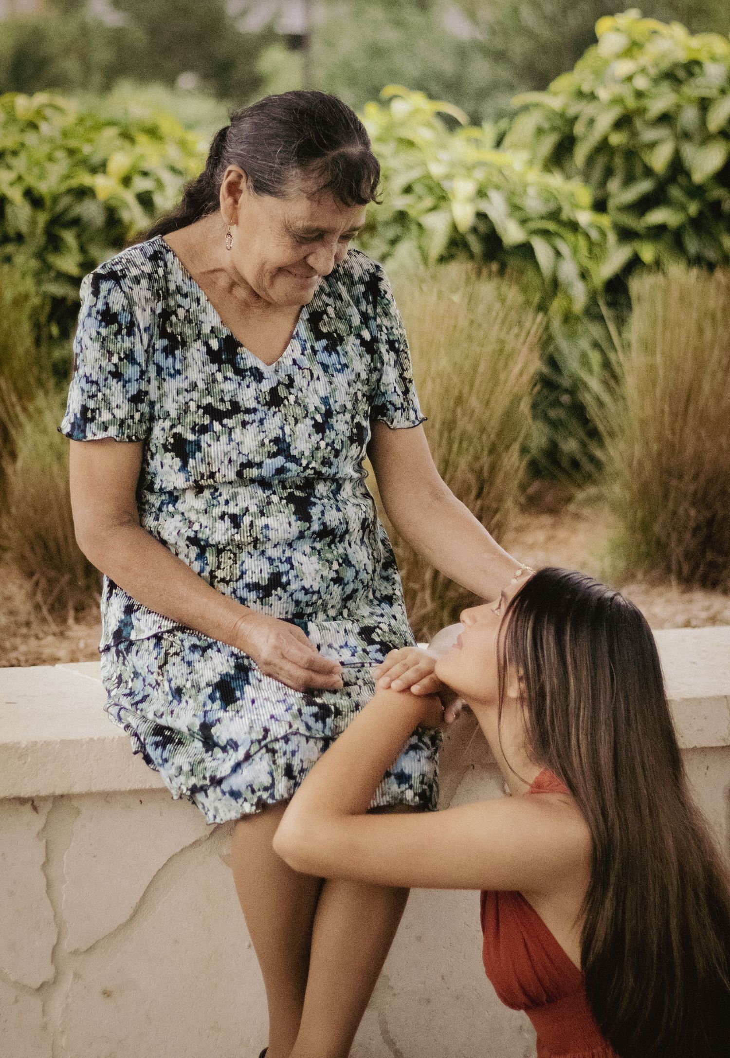 Elderly woman in floral dress tenderly stroking young girl's hair outdoors in a garden setting.