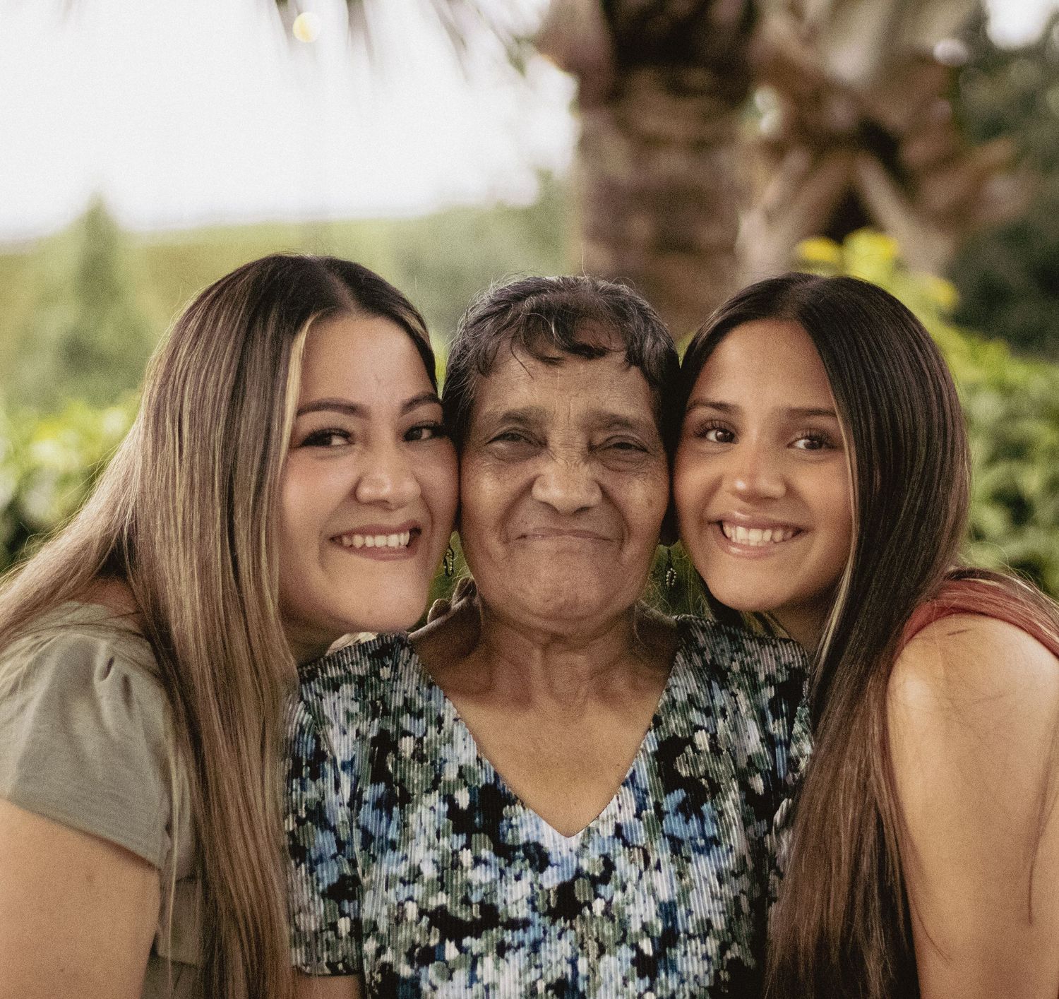 Elderly grandmother smiling warmly between two young women with long hair in an outdoor portrait.
