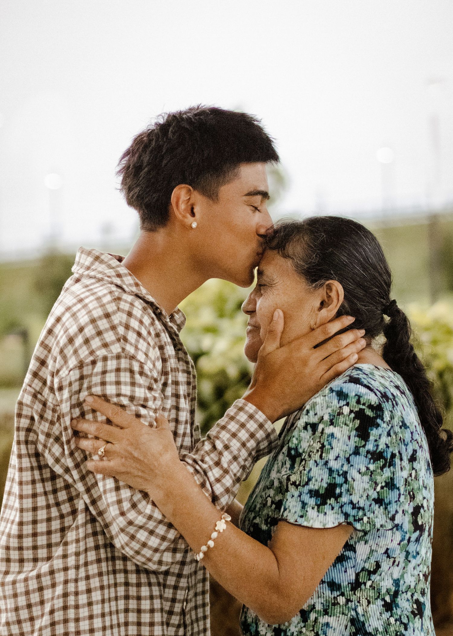 Young man tenderly kissing elderly woman on forehead while gently holding her face outdoors.