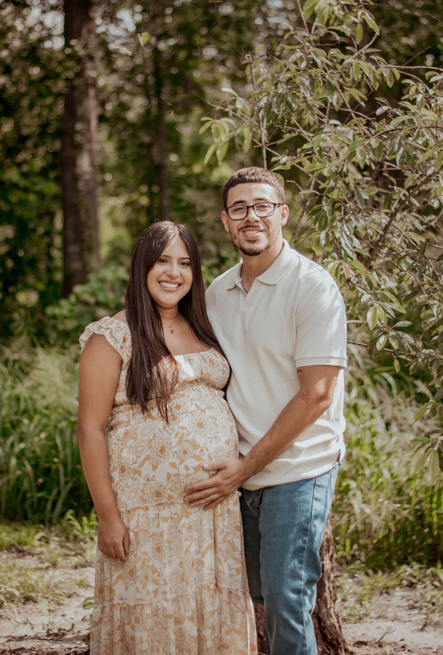 Smiling pregnant couple posing outdoors in a lush green forest setting during a maternity photo session.