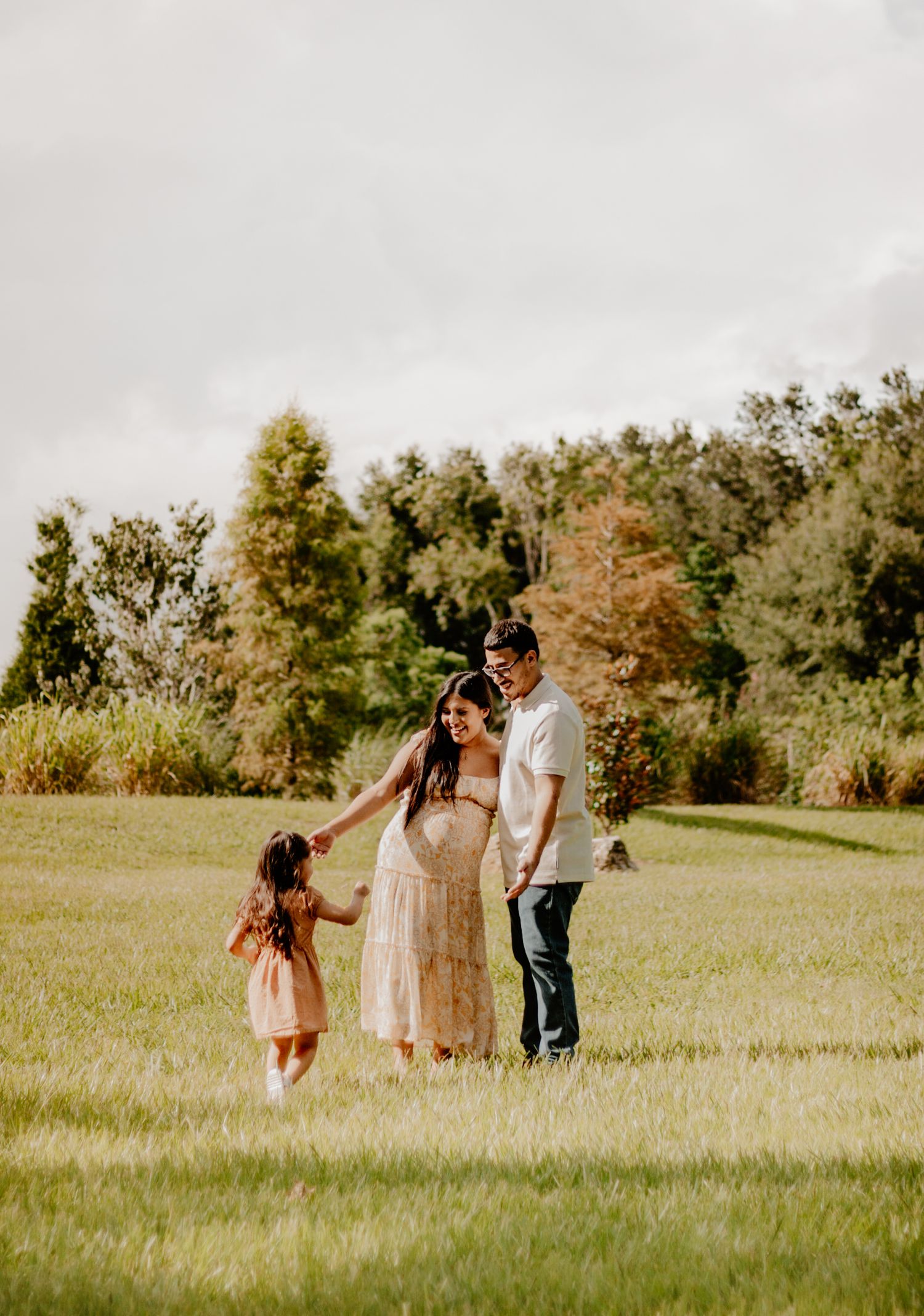 Pregnant woman and partner walking hand in hand with their young daughter across a green meadow.