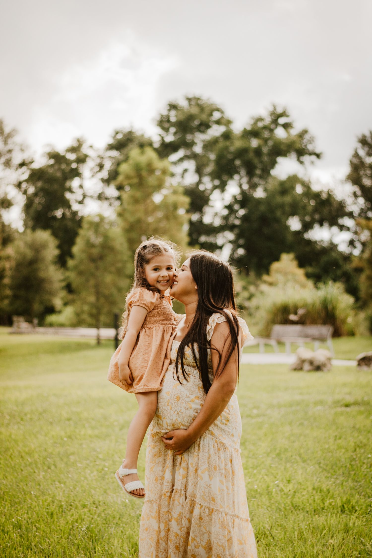 Pregnant mother kissing her smiling young daughter while standing in a lush green outdoor setting.