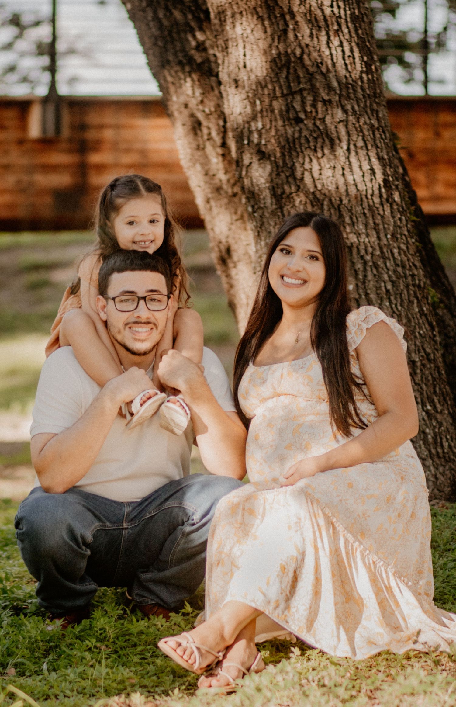 Family portrait of couple with young daughter on dad's shoulders sitting under a tree in golden hour light.