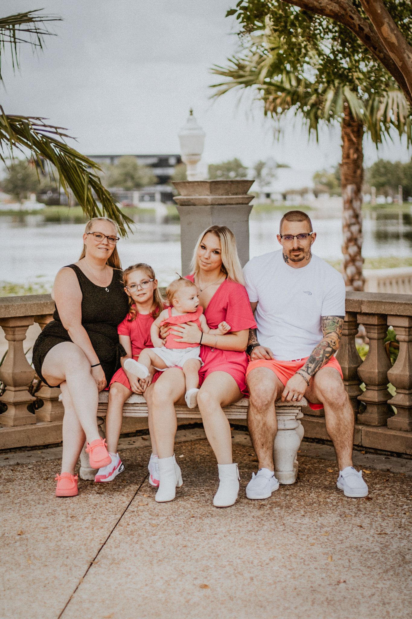 Family of five sitting on bench near waterfront, wearing coordinated pink and white outfits outdoors.
