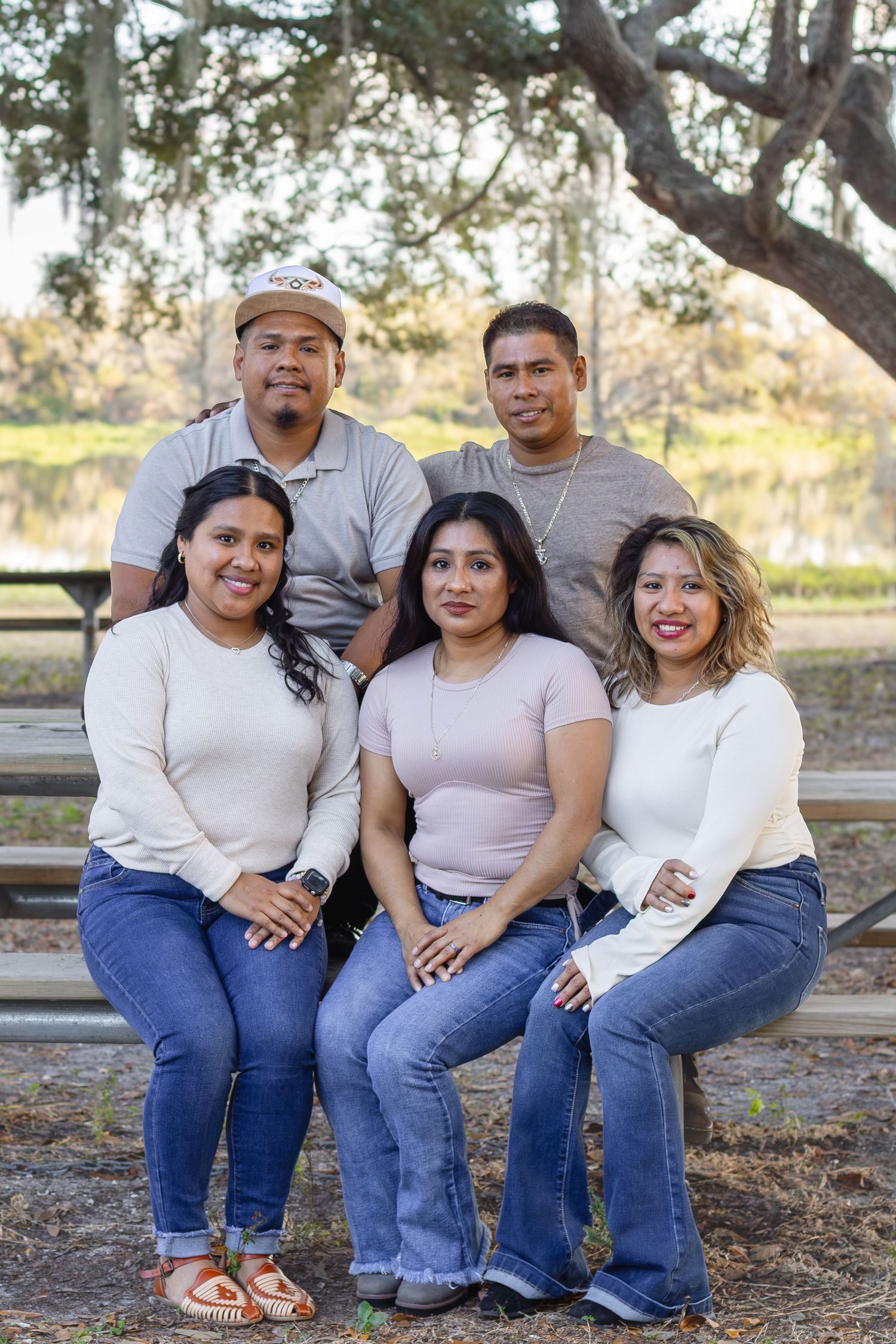 Five siblings posing together on a bench outdoors under a large tree in a park setting.