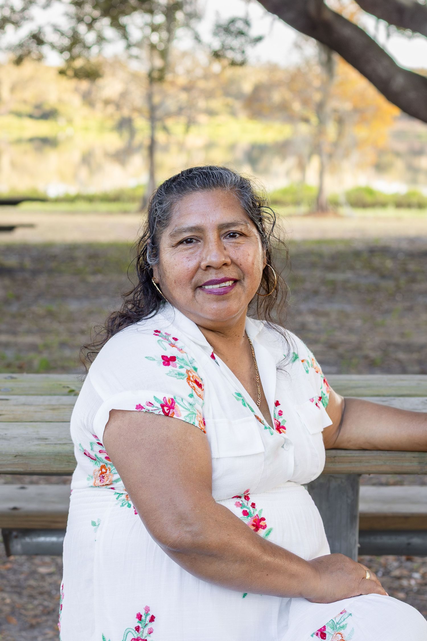 A smiling woman in a white floral dress sitting relaxed on a bench in an autumn park.
