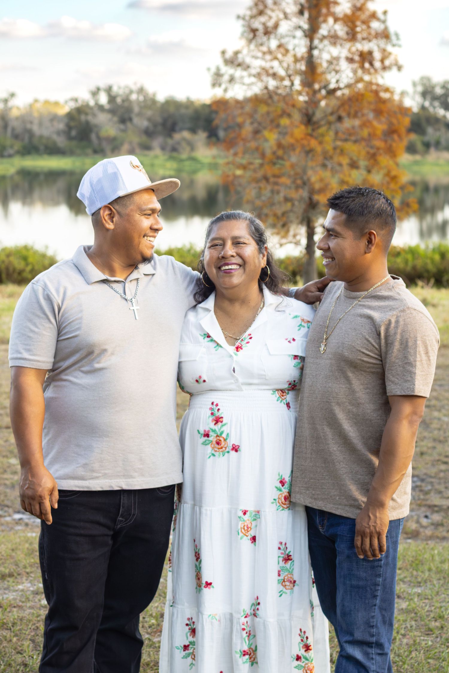 A woman in a floral dress laughing joyfully with two men beside a tranquil lake in autumn.