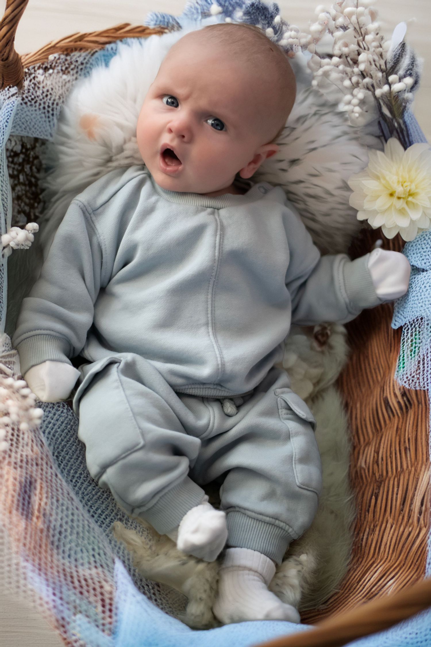 Baby in cozy light blue onesie lies on soft textured blanket with floral decorations.