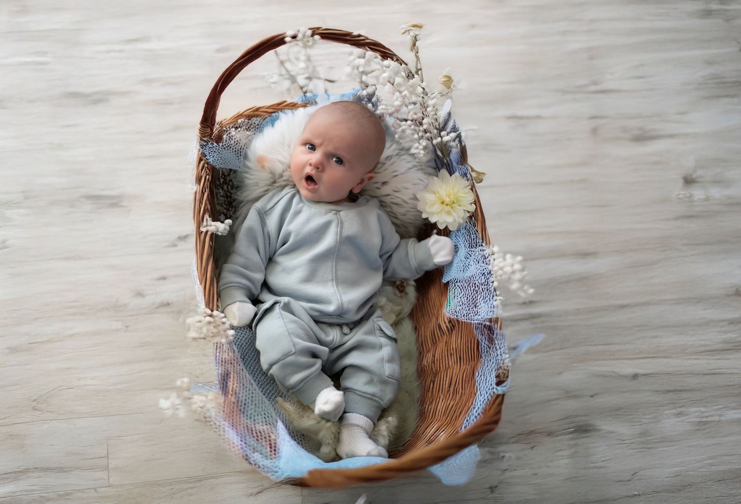 Baby rests in a brown wicker basket decorated with blue and white flowers on a light wooden floor.