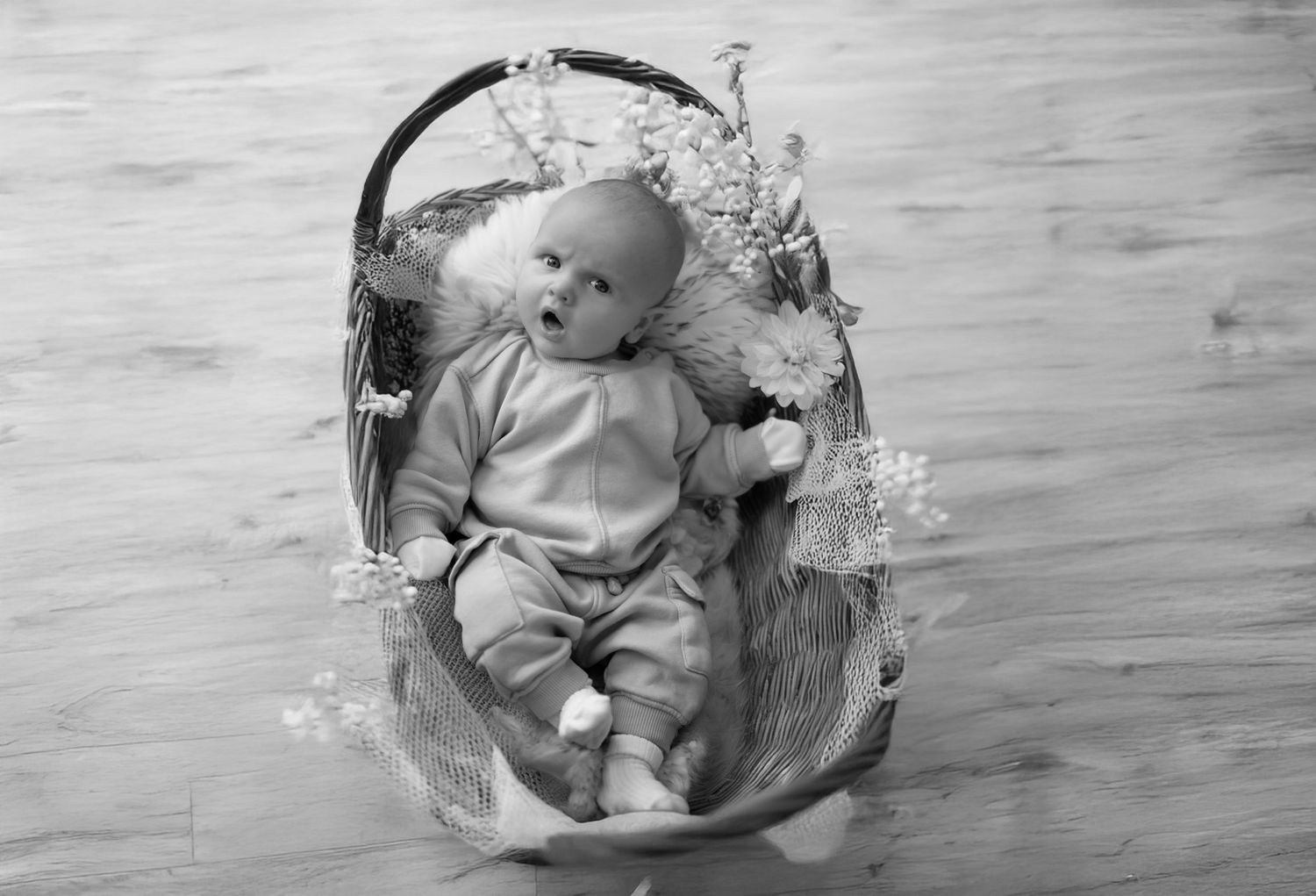 A small baby wearing gray clothes lies in a decorative basket surrounded by white flowers on a wooden floor.