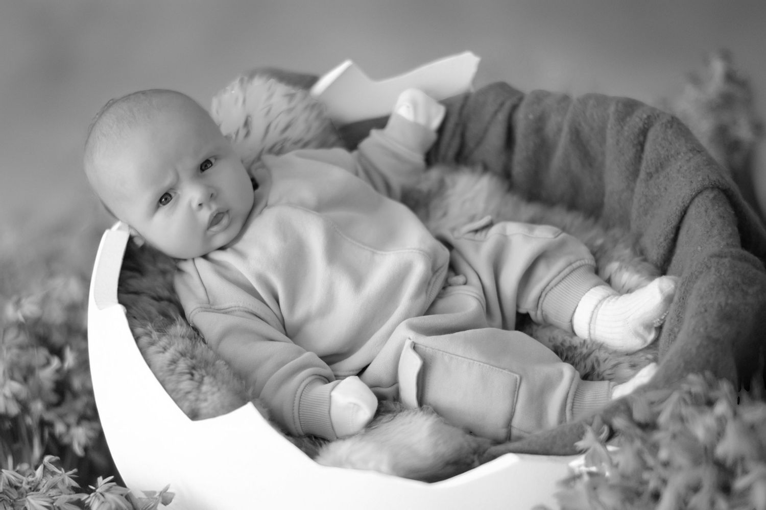 A baby in a light onesie lying contentedly in a crescent-shaped chair surrounded by flowers, captured in black and white.