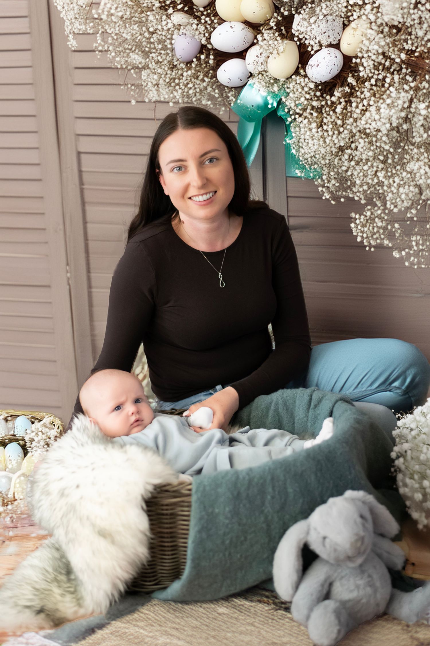 A mother and baby sit together in a cozy corner decorated with baby's breath flowers, pillows and a plush bunny toy.
