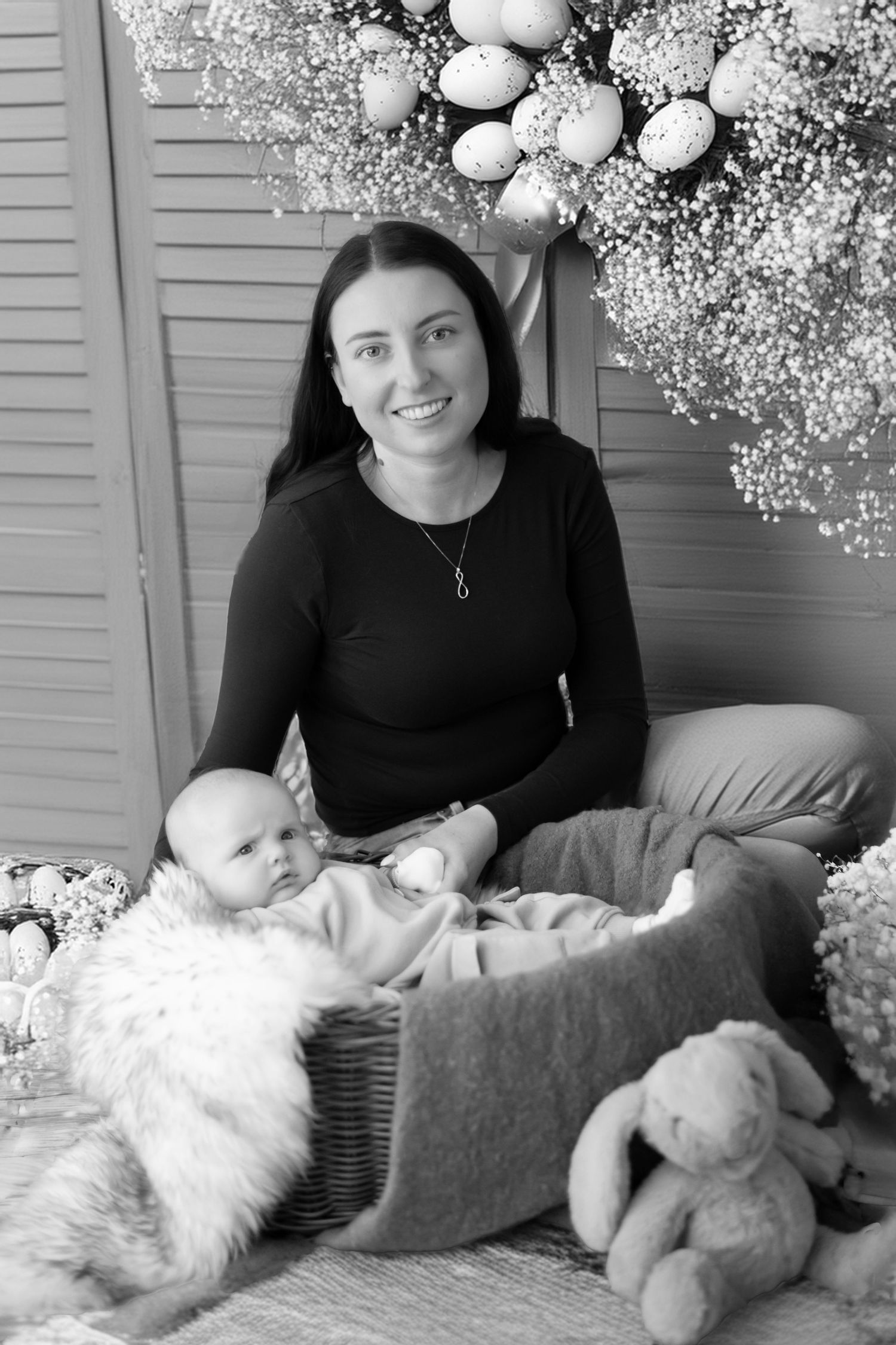 Black and white portrait of a parent with baby surrounded by soft plush toys and baby's breath flowers.