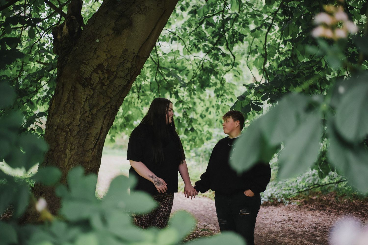 Two people in dark clothing walking along a tree-lined path surrounded by lush green foliage on a calm day.