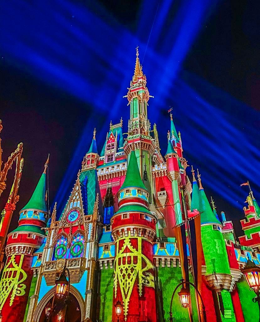 Cinderella Castle illuminated in festive green and red lights with dramatic blue spotlights.