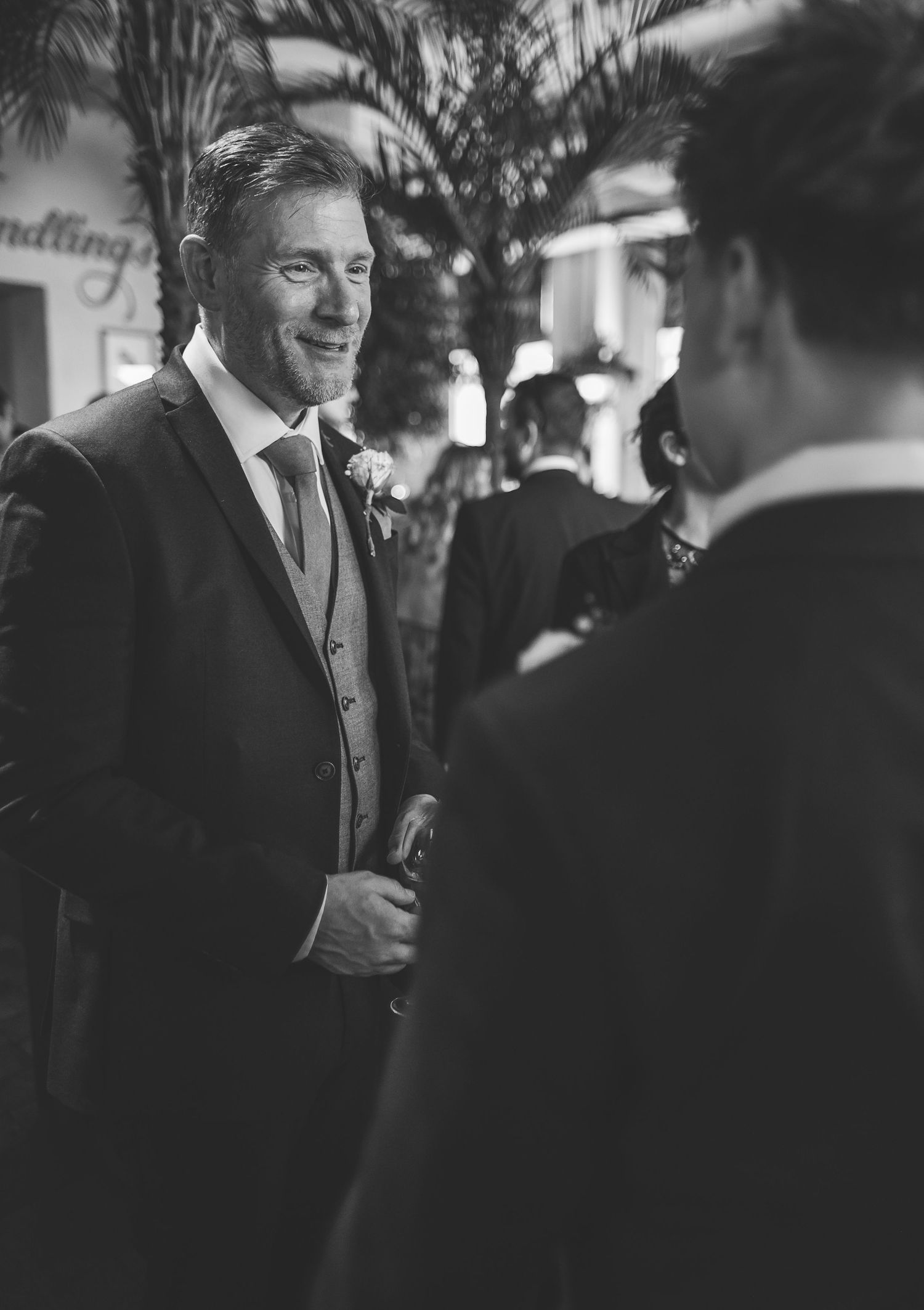 A well-dressed groom in a suit and tie engages in conversation at an elegant indoor reception drinks.