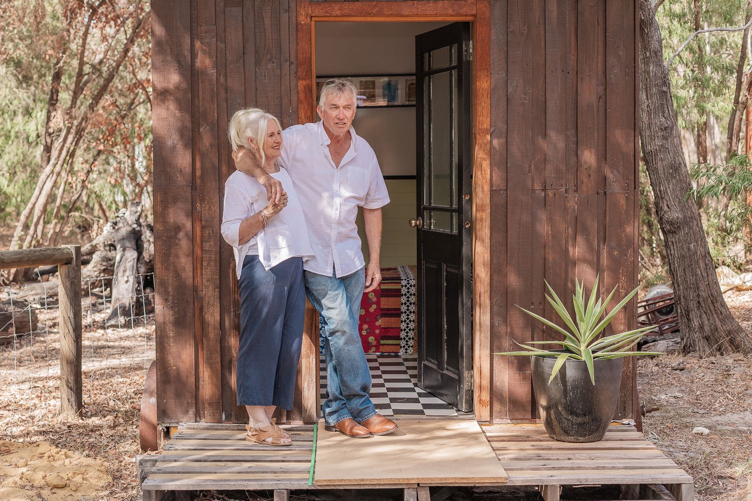 A couple stands in the doorway of a rustic wooden cabin with a potted plant on the deck.