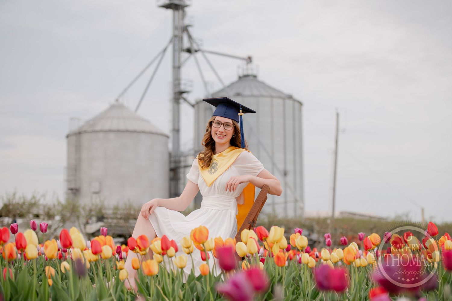 Someone in a graduation cap and gown sits in a vibrant field of colorful tulips with grain silos in the background.