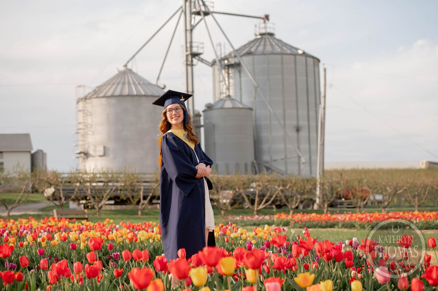 Graduate in cap and gown stands in vibrant red tulip field with grain silos in background.