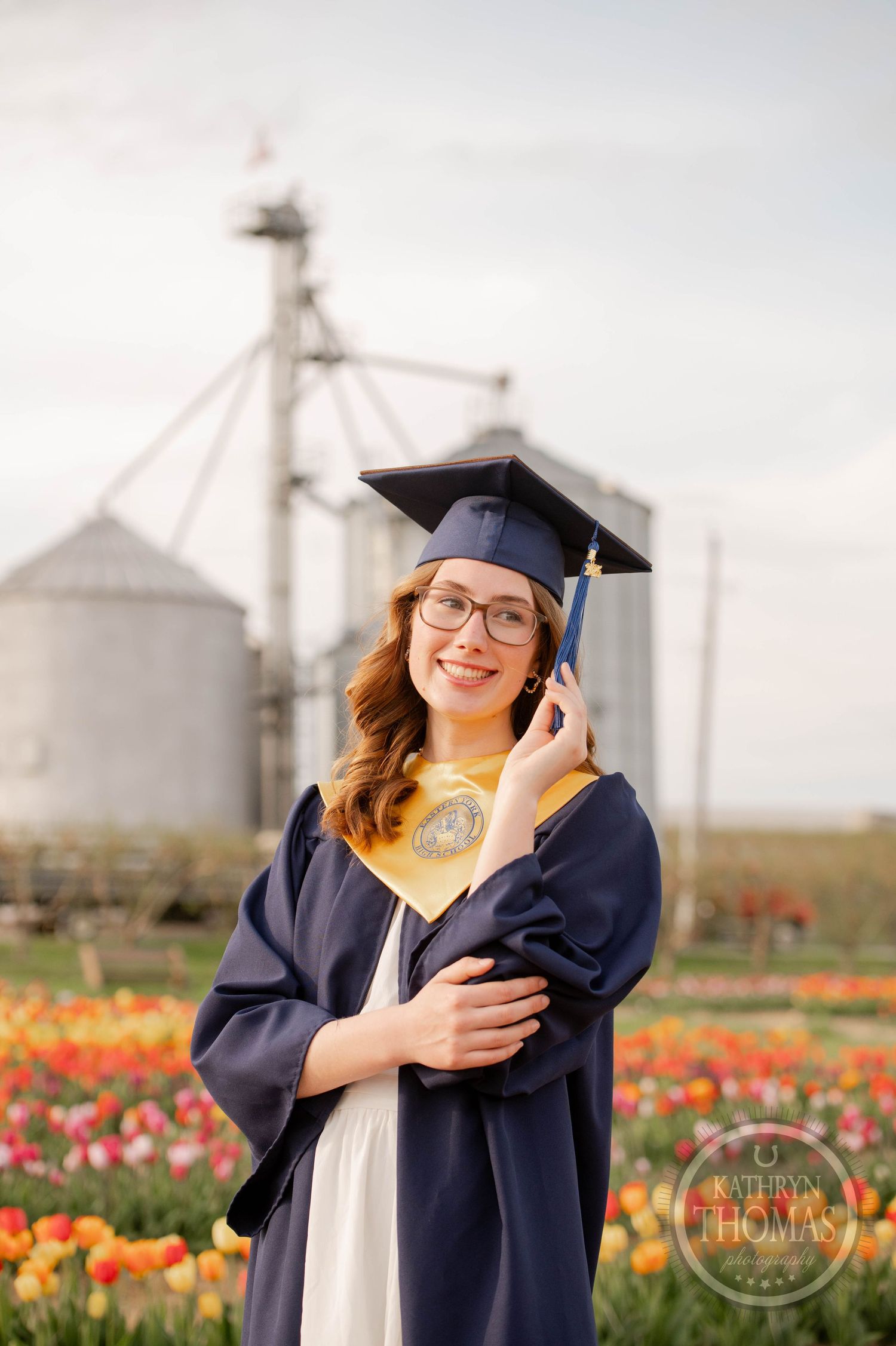 Graduate in cap and gown poses in front of grain silos surrounded by blooming tulip fields at sunset.