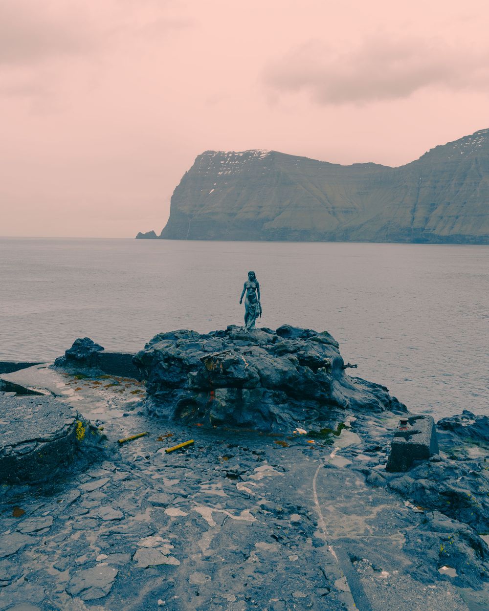 A silhouette stands on rocky coastal cliffs overlooking misty mountains and ocean during sunset creating a dramatic scene.