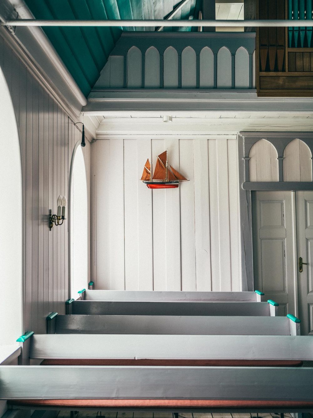 A model sailboat hangs on a white paneled wall above church-style pews with teal accents in a bright interior space.