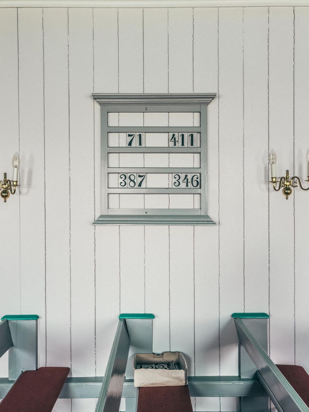 A white church interior wall features a blue-framed hymn board with numbers and three turquoise-trimmed wooden pews below.