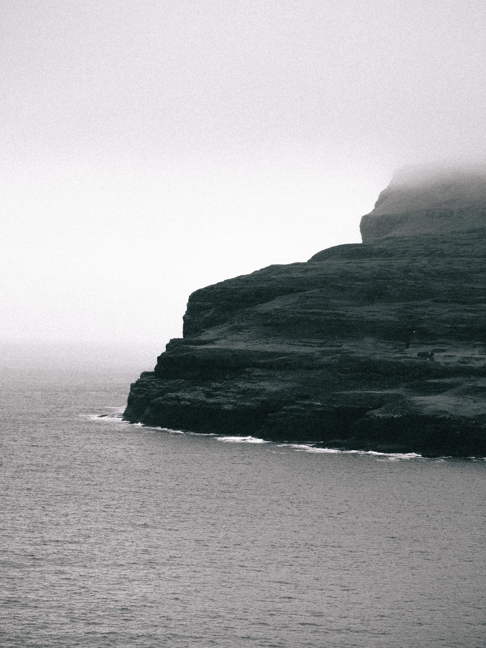 Dramatic black and white coastal cliffs shrouded in mist rise from the dark ocean waters below.