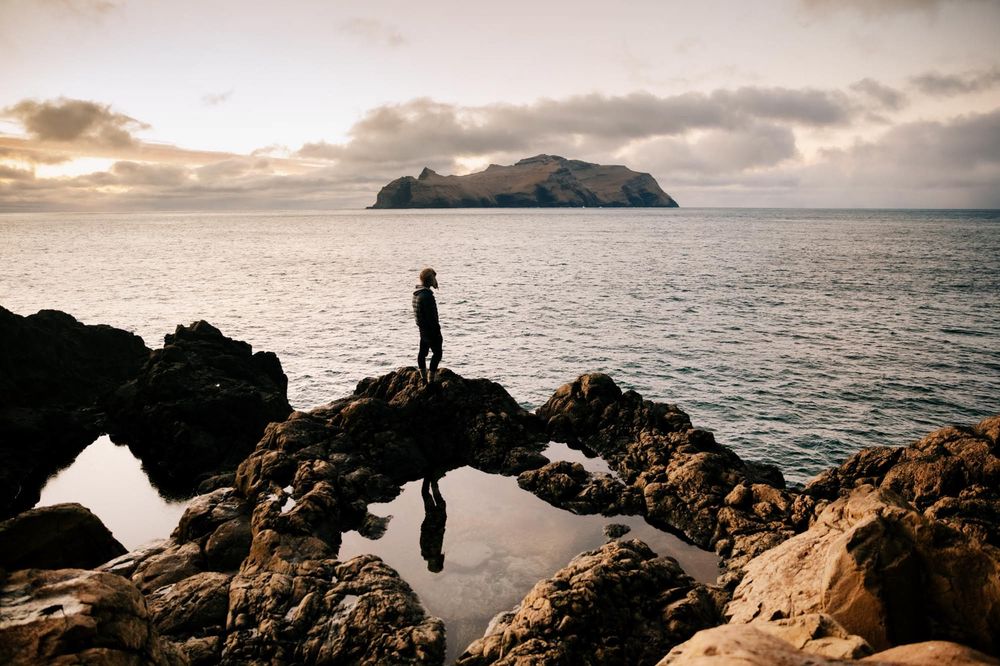 A silhouetted figure stands on coastal rocks overlooking a tranquil ocean with a distant island during golden sunset.