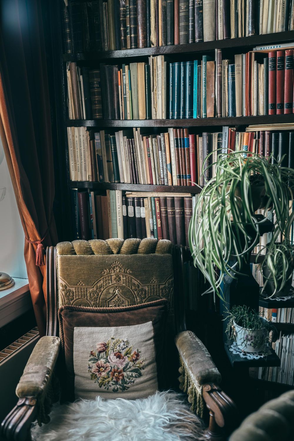 Vintage armchair with floral cushion sits beside bookshelf and potted plant in cozy reading nook.