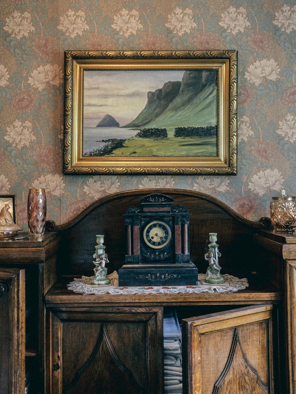 A vintage landscape painting in gold frame hangs above an antique wooden cabinet with ornate clock and floral wallpaper backdrop.