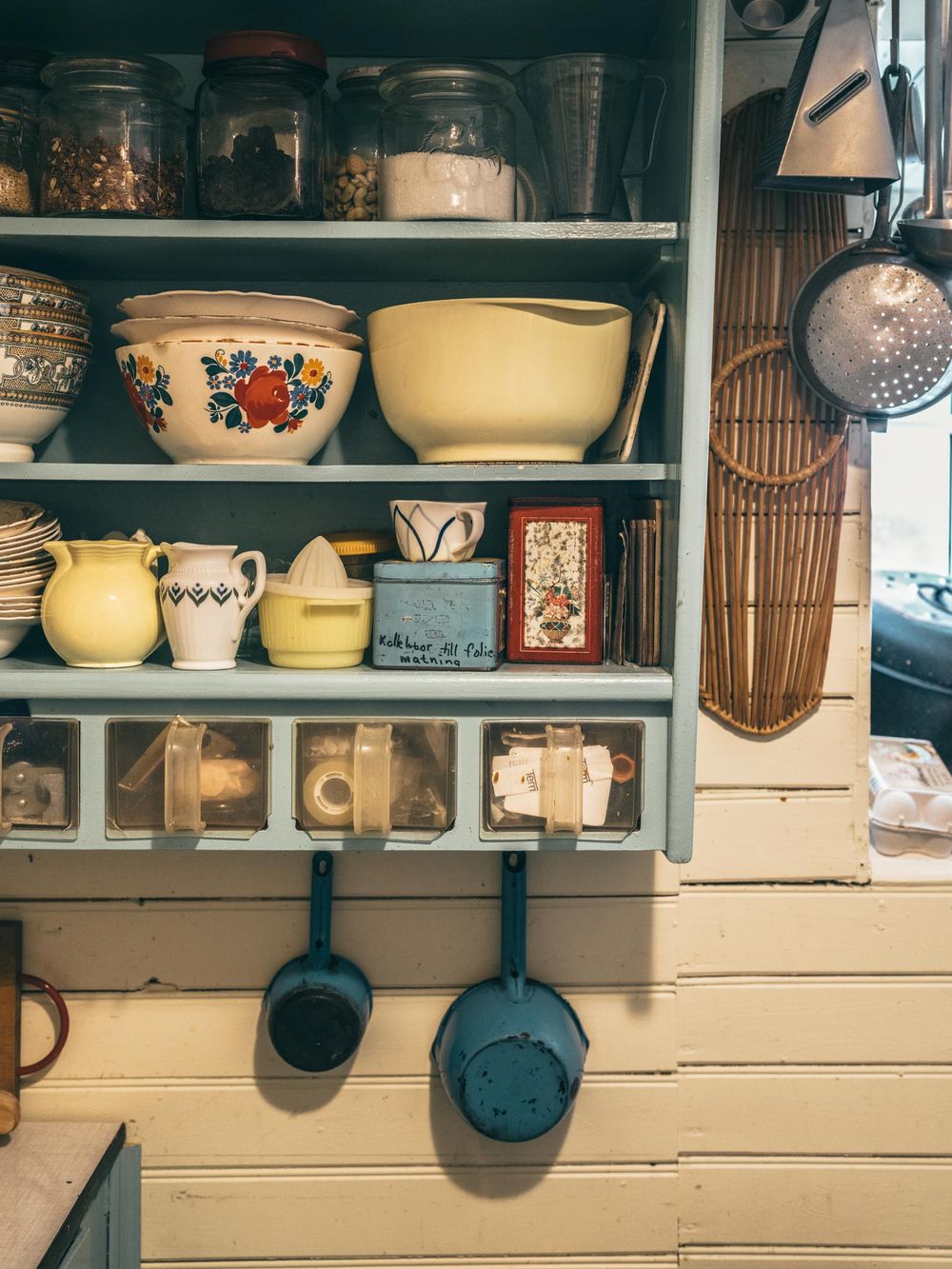 A vintage kitchen shelf displays colorful bowls, ceramic jars, and cast iron pans on a rustic painted wall.