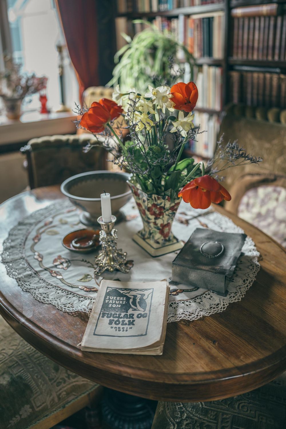 Vintage styled table setting with red tulips in vase, lace doily, and antique decor against bookshelf background.