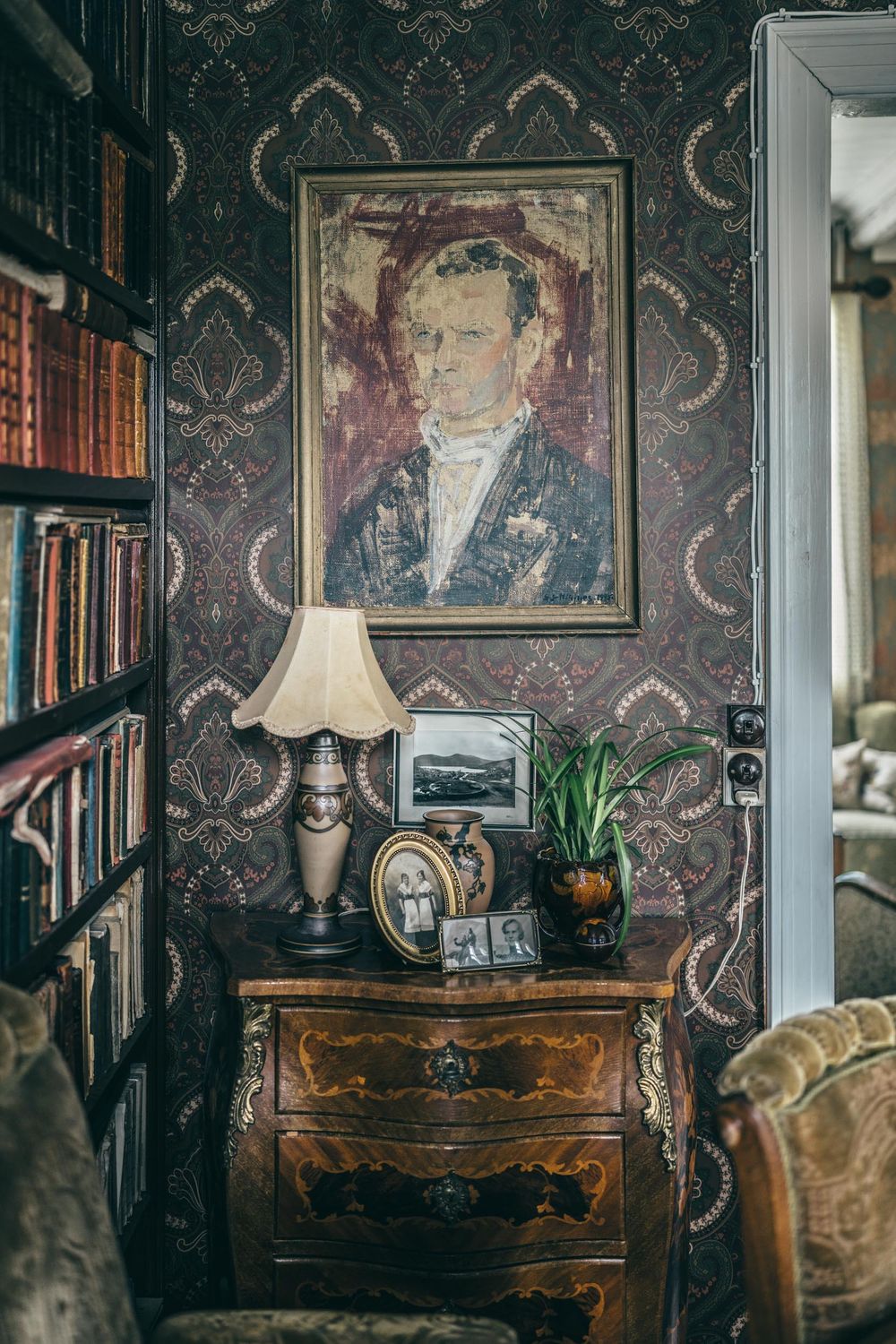 A vintage interior with ornate wallpaper, portrait painting, bookshelves, antique dresser, and table lamp atop it.