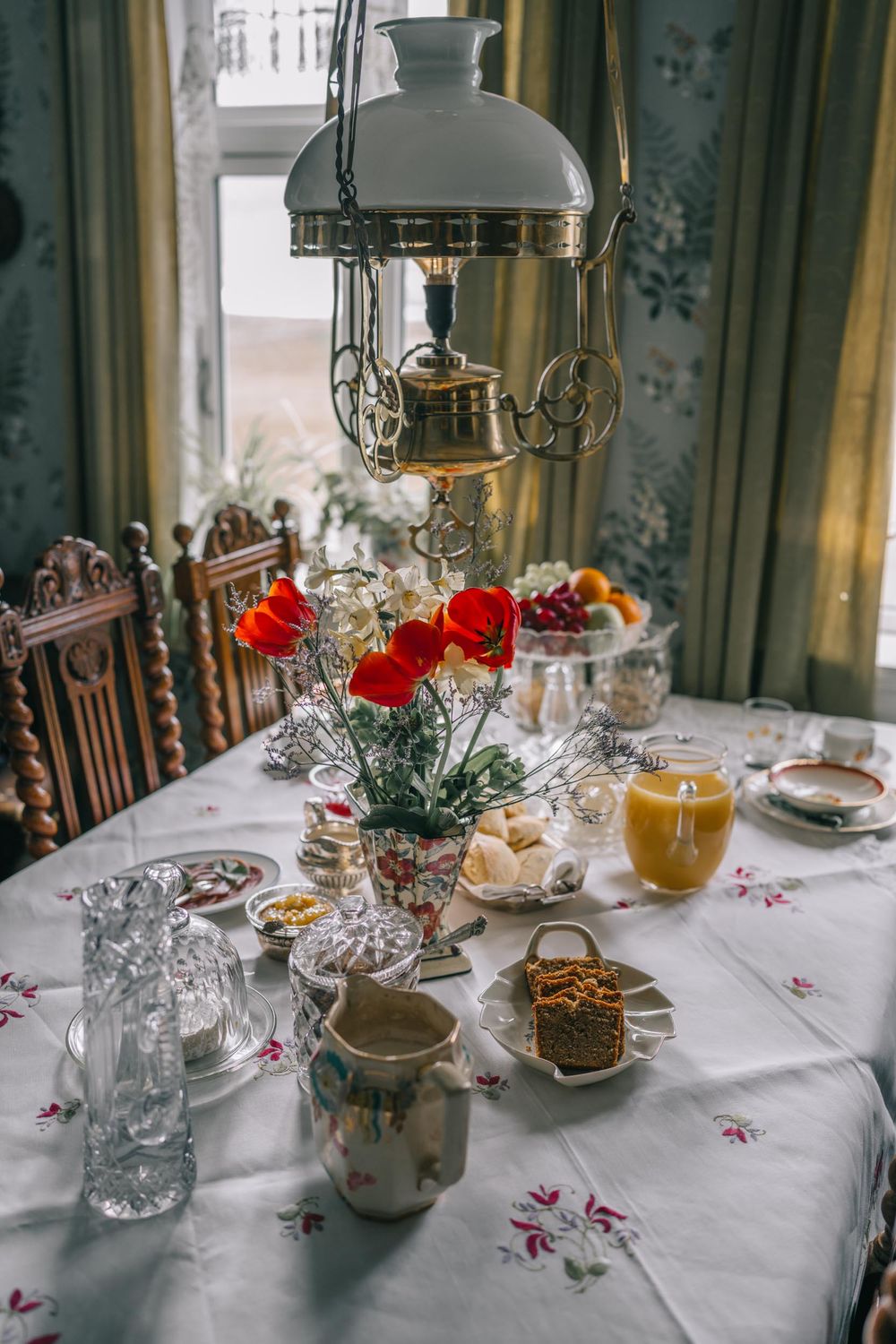 Vintage dining table set with ornate oil lamp, red poppies in crystal vase, and traditional breakfast settings on floral tablecloth.