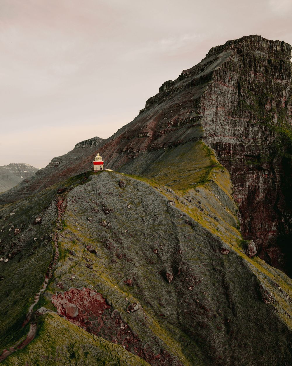 A small red lighthouse perches dramatically on a steep cliff face with moss-covered rocky terrain in the Faroe Islands.
