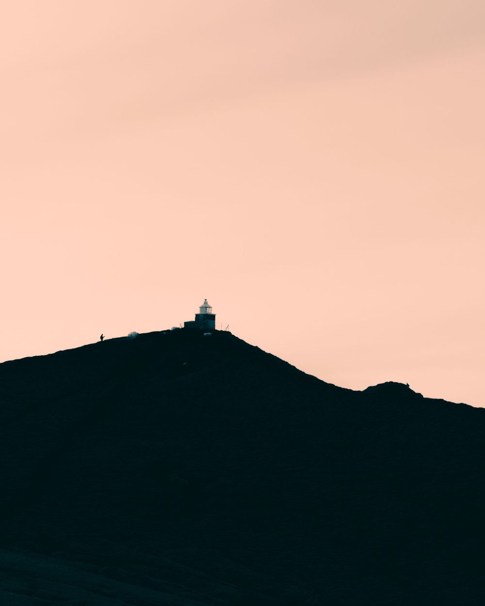 A silhouetted mountain ridge with a lighthouse tower at the peak against a warm pink sunset sky.
