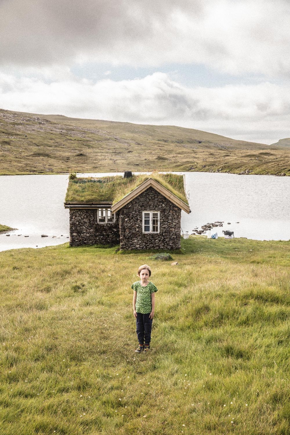 Traditional stone cottage with grass roof sits in remote countryside against snowy hills and cloudy sky in the Faroe Islands.