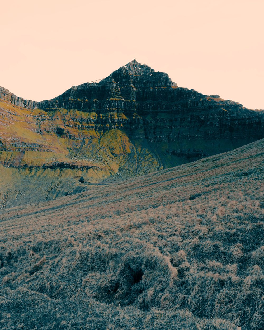 A rugged mountain peak rises against a pink sky with rocky slopes and scrubby vegetation in the foreground.