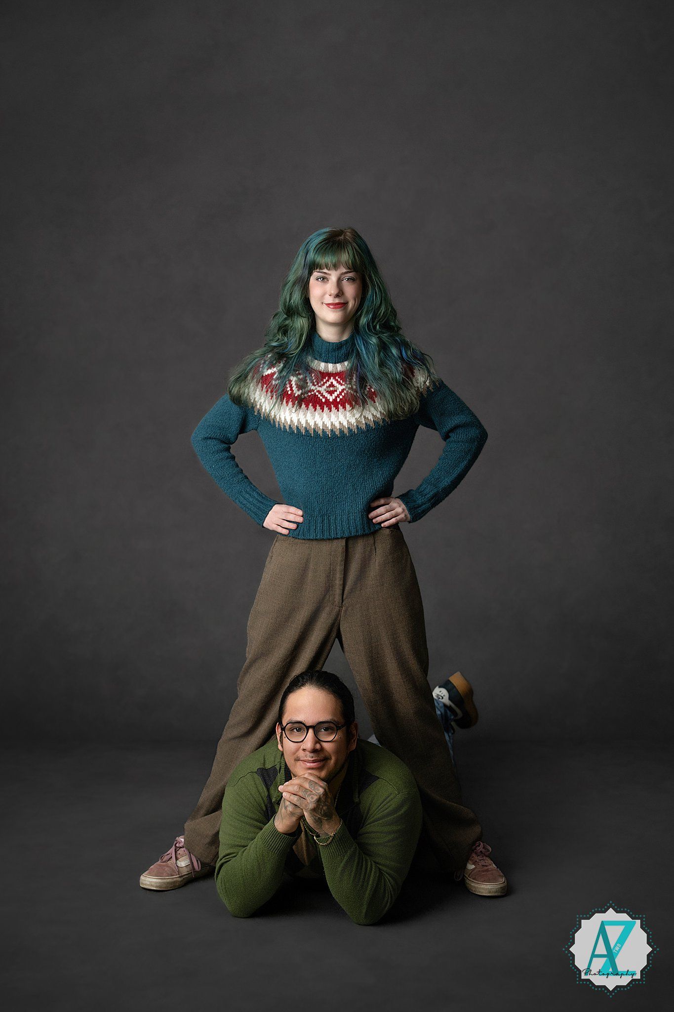 Two people pose creatively in a studio setting wearing winter sweaters against a dark grey backdrop.