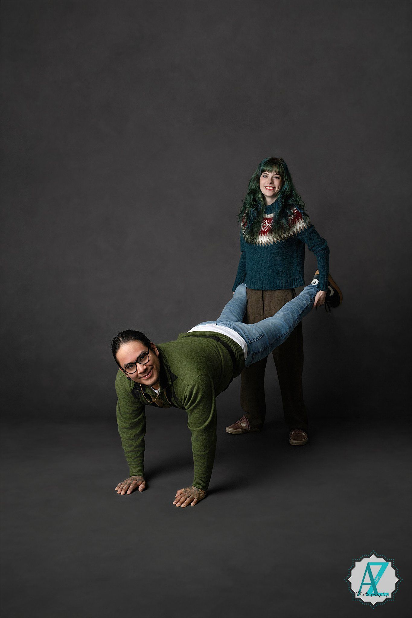 Two people pose playfully against a dark studio backdrop, one leaning forward while the other stands behind.