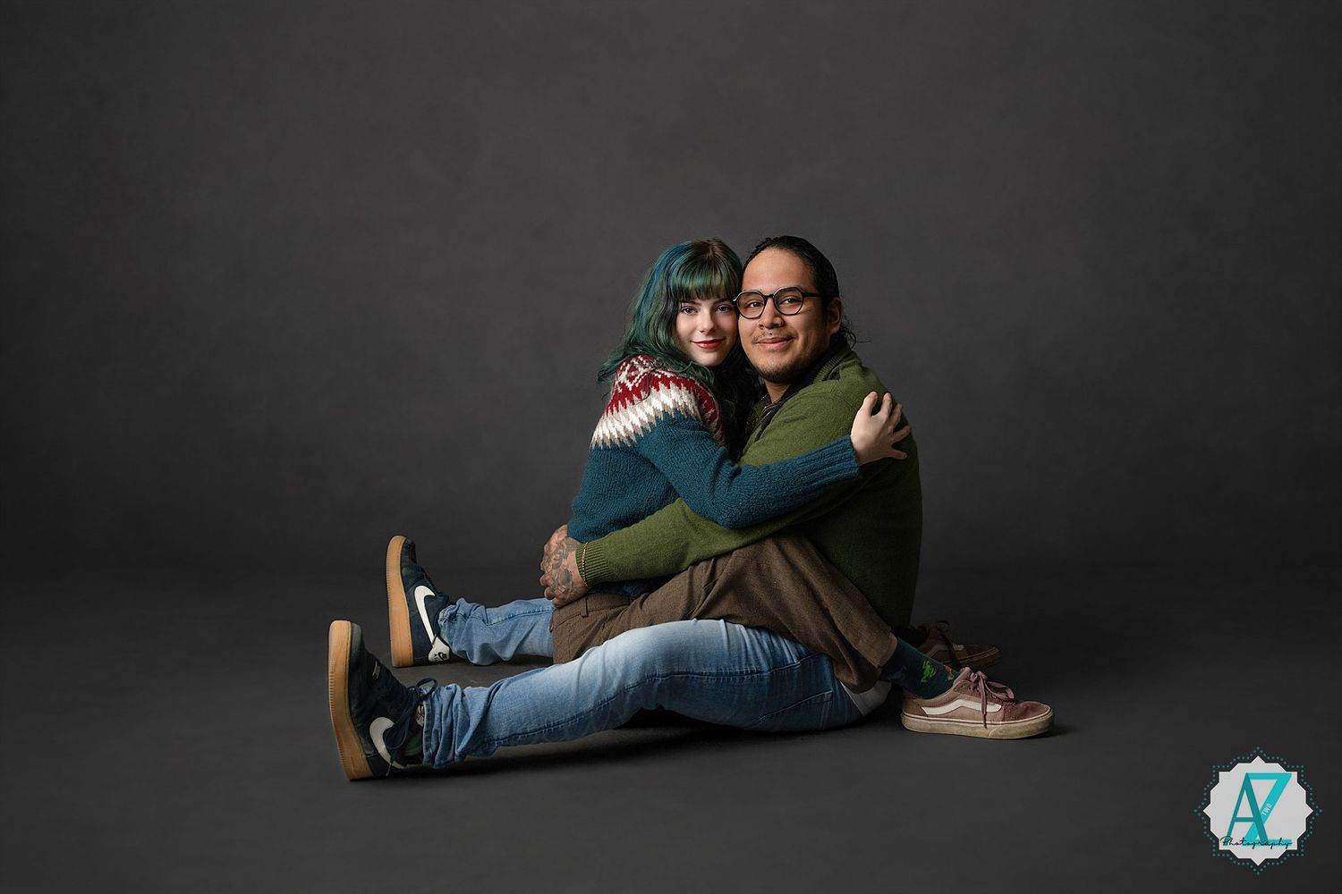 Two people sitting on the floor hugging and smiling against a dark gray studio backdrop.