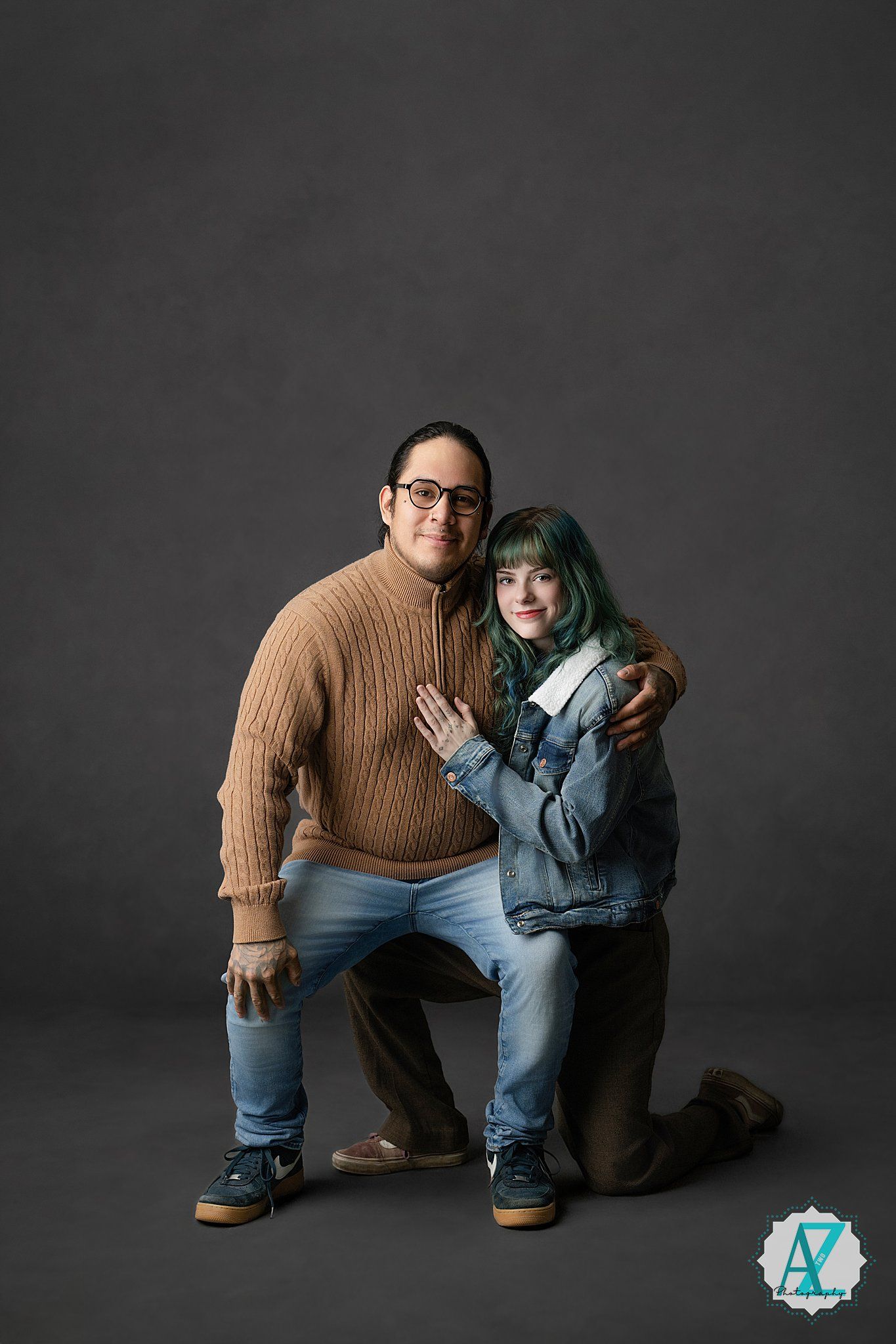 A couple in casual denim and sweater outfits poses against a dark backdrop in a studio portrait setting.