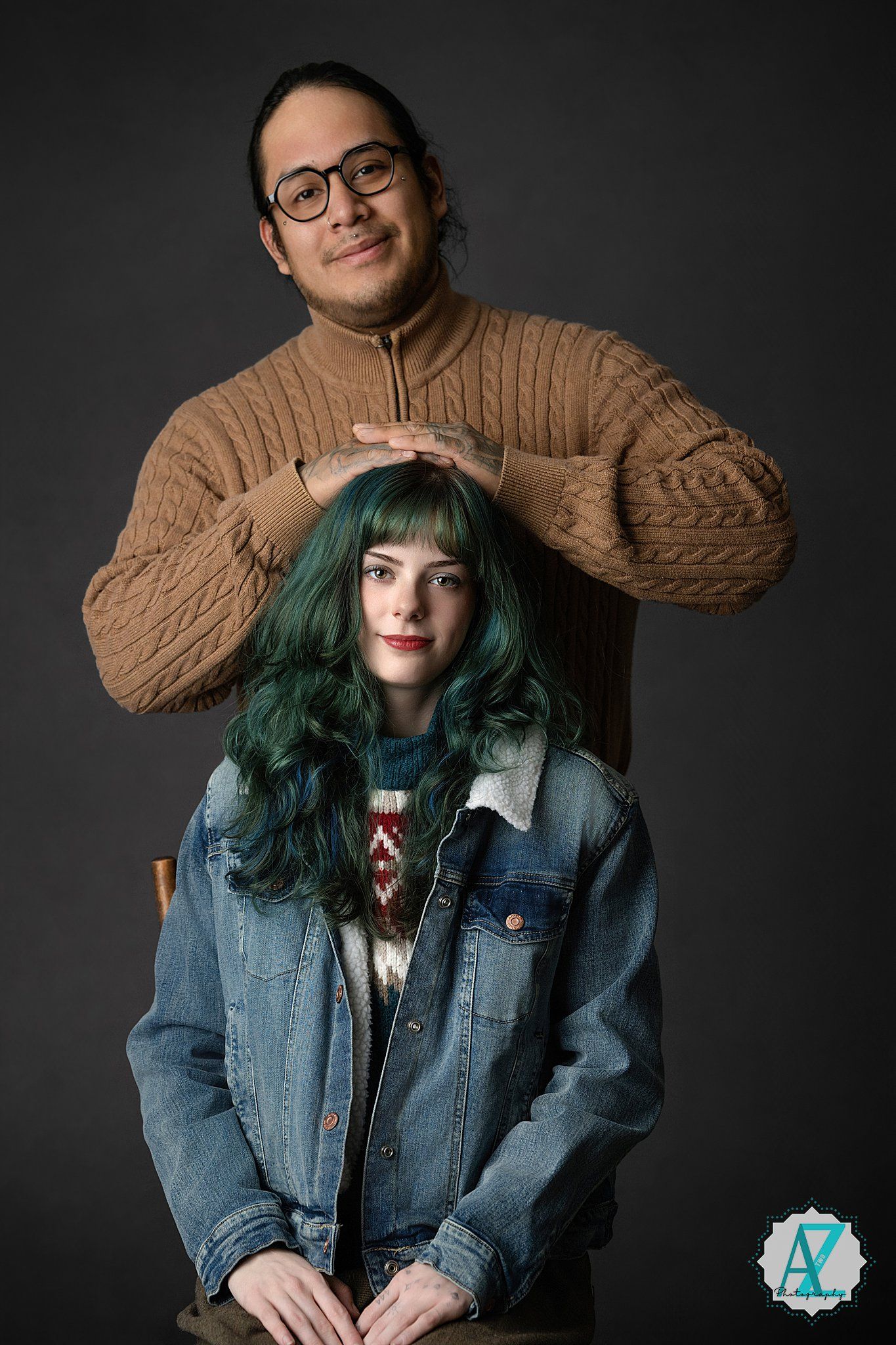 Two people pose playfully in casual clothing, with green hair and brown sweater creating visual contrast.