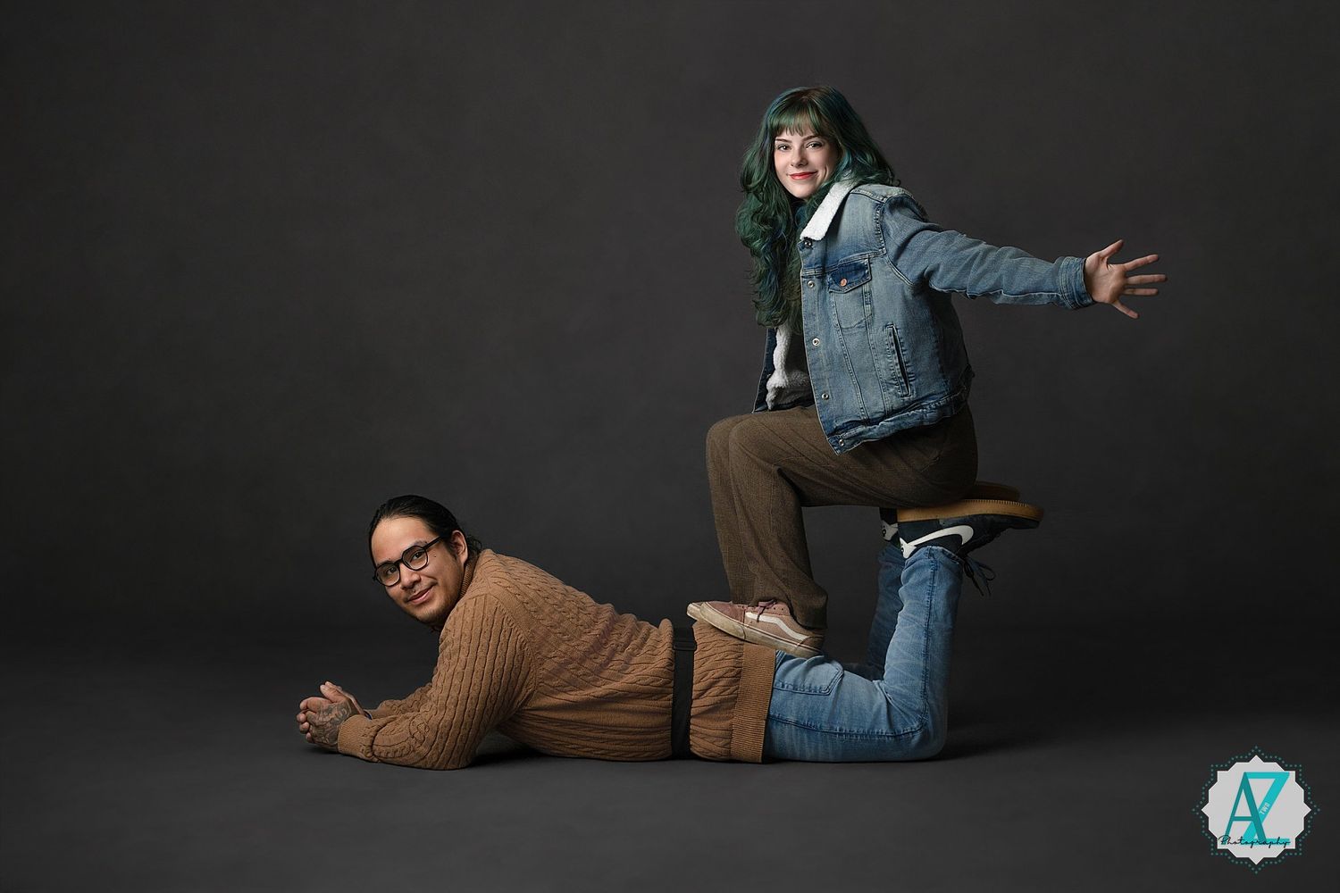 Two men pose playfully against dark backdrop, one lying down while other leaps over him in denim jacket.